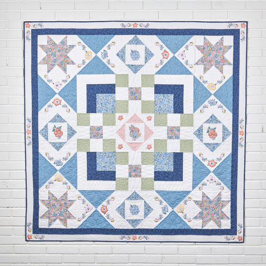 Colorful patchwork quilt with star and floral blocks in blue, white, green and pink tones, hanging on a white brick wall for display. image number