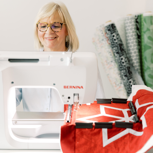 Smiling woman using a BERNINA embroidery machine to stitch a red quilt with a white star design, with neatly rolled fabrics in the background. image number
