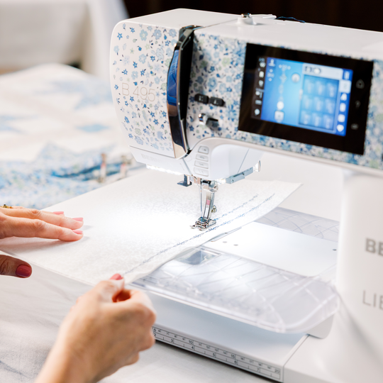 BERNINA sewing machine with blue floral design in use, stitching a white quilt sandwich under the needle with hands guiding the fabric. image number