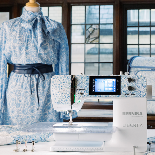 BERNINA LIBERTY sewing machine on a table with presser feet, shown in front of a dress form wearing a blue printed dress in a bright studio. image number
