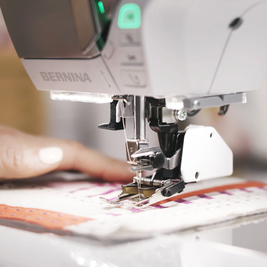 BERNINA N&auml;hmaschine B 570 QE beim Quilten, Nahaufnahme von N&auml;hfuss und Transporteur, eine Hand f&uuml;hrt mehrere bunte Stofflagen image number