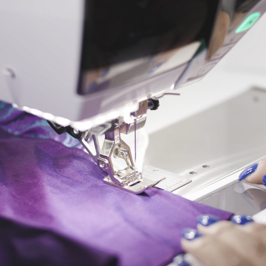 Person n&auml;ht lila Stoff an einer BERNINA N&auml;hmaschine, Nahaufnahme vom N&auml;hfuss mit transportiertem Material und H&auml;nden am Stoff image number
