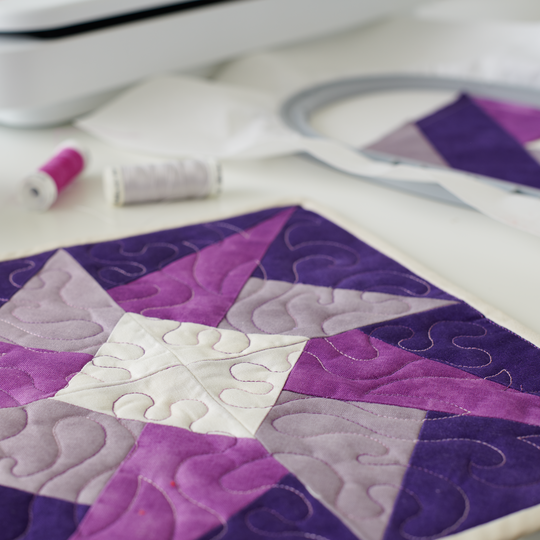 Lila Patchwork-Quiltblock mit weissem Quiltmuster liegt neben Stickrahmen, Garnspulen und einer teilweise sichtbaren BERNINA Stickmaschine image number