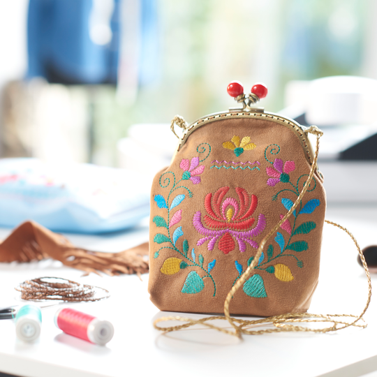 Kleine braune Tasche mit farbenfrohem Blumenstickmotiv und Kugelverschluss steht auf einem Tisch, umgeben von Garnrollen und weiterem N&auml;hzubeh&ouml;r image number