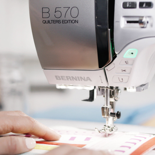 BERNINA B 570 QE N&auml;hmaschine in Nahaufnahme beim Quilten, mit sichtbarem N&auml;hfuss, leuchtenden Tasten und H&auml;nden, die einen Quilt f&uuml;hren image number