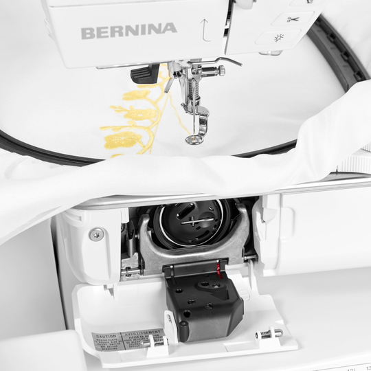 BERNINA N&auml;hmaschine mit ge&ouml;ffneter Greiferabdeckung, sichtbar eingesetzter Spule und Stoff im Stickrahmen unter dem N&auml;hfuss image number