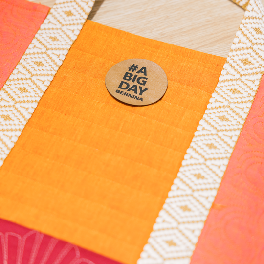 Nahaufnahme einer gen&auml;hten Stofftasche in Orange und Pink mit gemusterten Henkeln und rundem BERNINA Badge mit der Aufschrift &bdquo;A BIG DAY&ldquo;. image number
