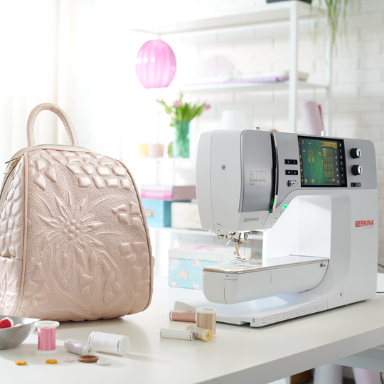 BERNINA 770 PLUS in heller Nahecke, mit grossem Touchscreen; daneben eine gesteppte beigefarbene Tasche und verstreute N&auml;hutensilien image number