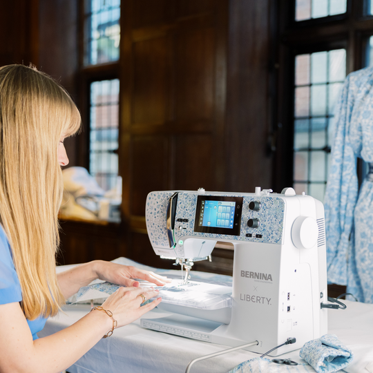 Woman sewing on the BERNINA 5 Series LIBERTY sewing and embroidery machine in a bright room, with a blue patterned dress displayed in the background. image number