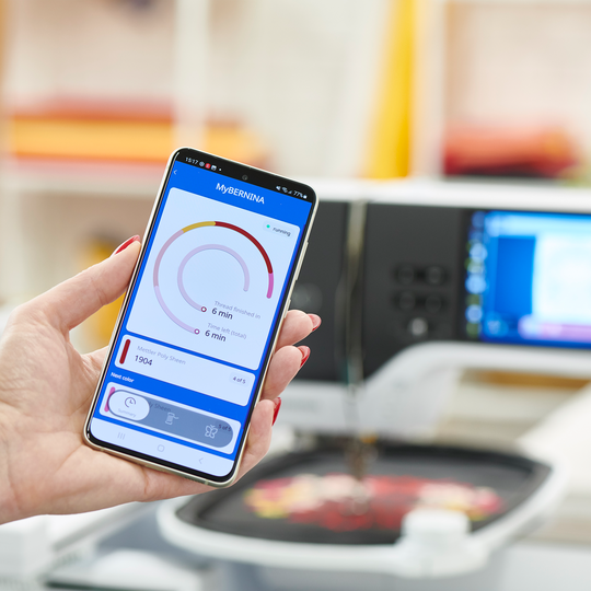 Hand haelt Smartphone mit geoeffneter BERNINA App vor einer BERNINA 790 PRO Stickmaschine, die ein buntes Motiv in einem Stickrahmen stickt image number