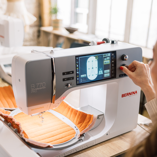 Person bedient die BERNINA 770 QE PLUS im Stickmodus, ein orangestoff gespannter Stickrahmen liegt unter der Nadel in heller Atelierumgebung image number