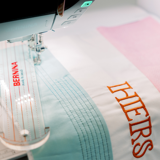 BERNINA sewing machine quilting straight parallel lines on white fabric with a clear BERNINA marked ruler next to orange &ldquo;HERS&rdquo; embroidery text. image number