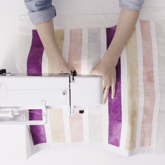 Person quiltet mit einer BERNINA 570 QE eine bunte Decke mit violetten, rosa und beigen Streifen, Ansicht von oben auf Maschine und Stoff image number