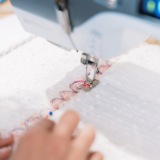 Hand fuehrt weissen Stoff mit farbiger Ziernaht unter dem N&auml;hfuss einer BERNINA N&auml;hmaschine, Nahaufnahme der Nahtfuehrung image number