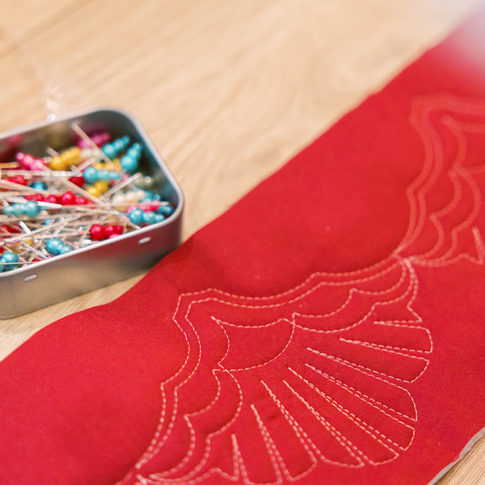 Rotes Quiltprojekt mit dekorativen, hellen Steppn&auml;hten in Muschelform auf Holzuntergrund, daneben eine ge&ouml;ffnete Dose mit bunten Stecknadeln image number