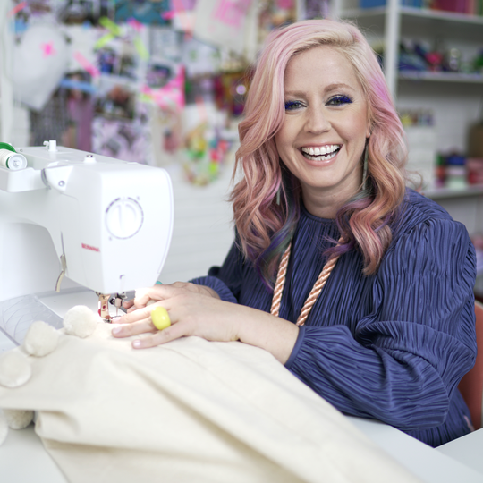 Lachende Frau mit rosa Haaren n&auml;ht einen hellen Stoff an einer weissen BERNINA N&auml;hmaschine in einem bunt eingerichteten N&auml;hatelier image number