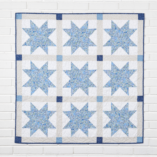 Light blue and white patchwork quilt with repeating star blocks, hanging flat against a white brick wall to show the full quilting pattern. image number